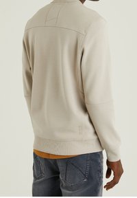 Svetle béžová mikina s žebrovanými manžetami a lemem, s texturovaným zadním panelem a jednoduchými švovými detaily. Nosená přes tmavě oranžovou košili.