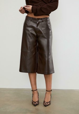 Femme portant des culottes en cuir marron foncé et des talons hauts imprimés léopard, debout sur un sol uni avec un mur blanc en arrière-plan.