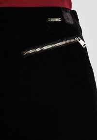 Zwarte fluwelen rok met een zijzak met ritssluiting en zilverkleurige hardware, en een klein gouden logo-tag op de tailleband.