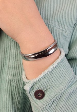 Metalen bangle armband met een gepolijste zilveren afwerking, gebogen ontwerp met een gestructureerd gaasgedeelte, gedragen om een pols met een zachte groene corduroy mouw.