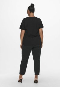 Black cotton t-shirt with short sleeves, relaxed fit. Worn with dark gray trousers and black heels, shown from the back against a plain background.