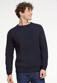 Marineblauer Kabelstrickpullover mit Rundhalsausschnitt, gerippten Säumen und Bündchen. Über einer blauen Jeans getragen, nutzt er ein strukturiertes Muster und eine taillierte Passform.