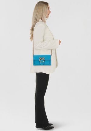 Borsa a tracolla in pelle blu con hardware dorato, design rettangolare e chiusura a pattina. Il modello indossa un blazer bianco e pantaloni neri.