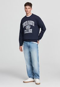Felpa della marina con testo e emblema bianchi "NORTH DAKOTA COLLEGE", abbinata a jeans azzurri chiari e sneakers bianche. Vestibilità casual, polsini a costine.