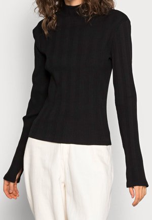 Žena v přiléhavém černém žebrovaném svetru s rolákem a dlouhými rukávy, v bílých kalhotách s vysokým pasem, na jednotném pozadí.