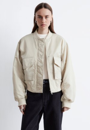 Ung kvinde iført en oversized beige bomberjakke med forlommer, hvid top, sorte bukser og sølvfarvede creoløreringe, stående mod en hvid baggrund.