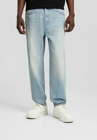 Vaqueros de mezclilla azul claro con un ajuste relajado, que presentan un ligero desvanecimiento, diseño clásico de cinco bolsillos y una textura desgastada. Combinados con zapatillas blancas.
