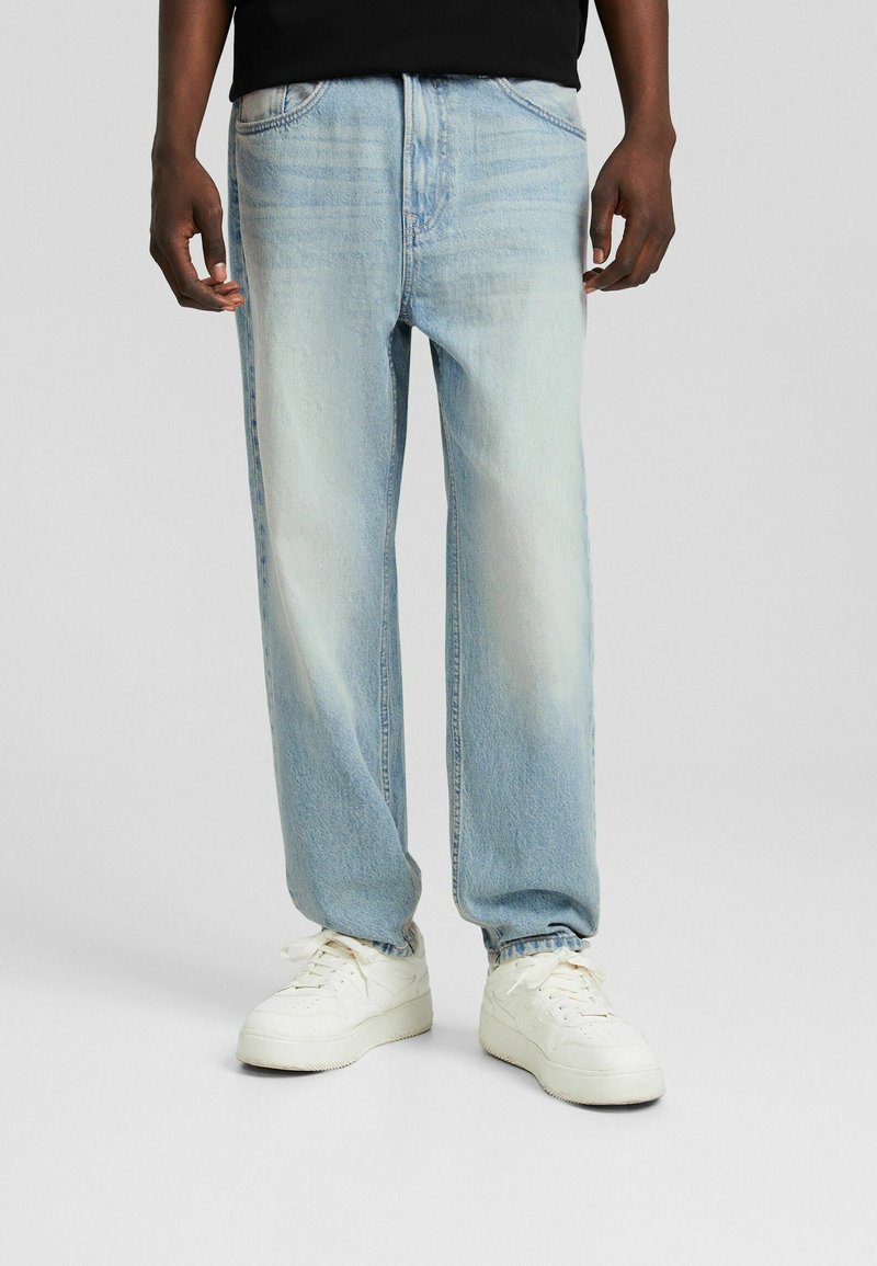 Vaqueros de mezclilla azul claro con un ajuste relajado, que presentan un ligero desvanecimiento, diseño clásico de cinco bolsillos y una textura desgastada. Combinados con zapatillas blancas.