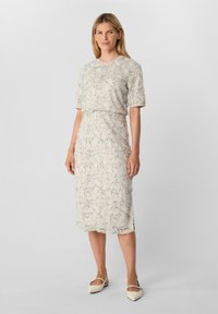 Femme portant une robe beige à mi-longueur avec des broderies florales et des manches jusqu'au coude, se tenant devant un fond clair uni.