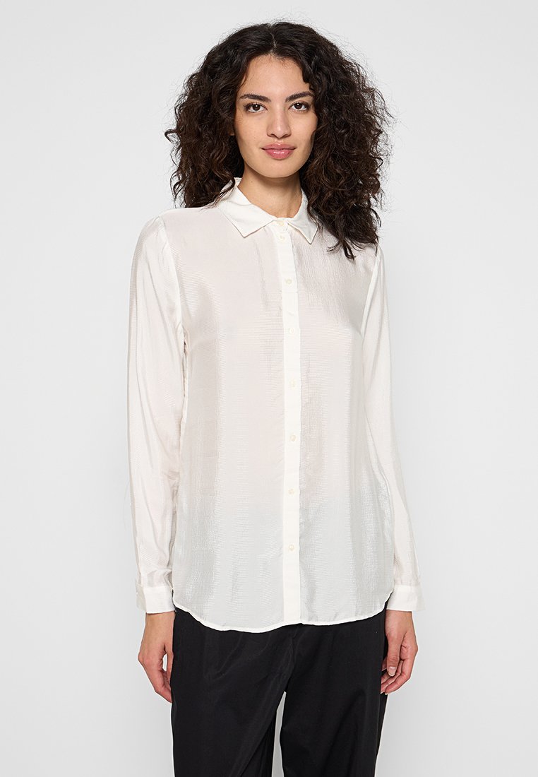 Mujer con cabello rizado que lleva una blusa blanca de botón de mangas largas y pantalones negros, de pie contra un fondo claro y liso.