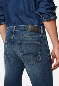 Dark blue denim jeans with a fitted design, featuring back pockets with curved seams and a black leather brand patch on the waistband.