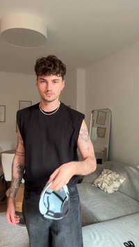 Camisa preta sem mangas, acessório azul segurado na mão, jeans escuros, tatuagens visíveis nos braços, sofá de cor clara ao fundo. Decoração minimalista.