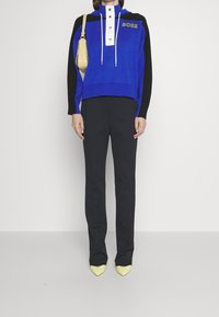 Person klädd i en blå och svart BOSS-tröja, svarta byxor, gula spetsiga skor och bär en liten beigemelerad handväska på axeln.