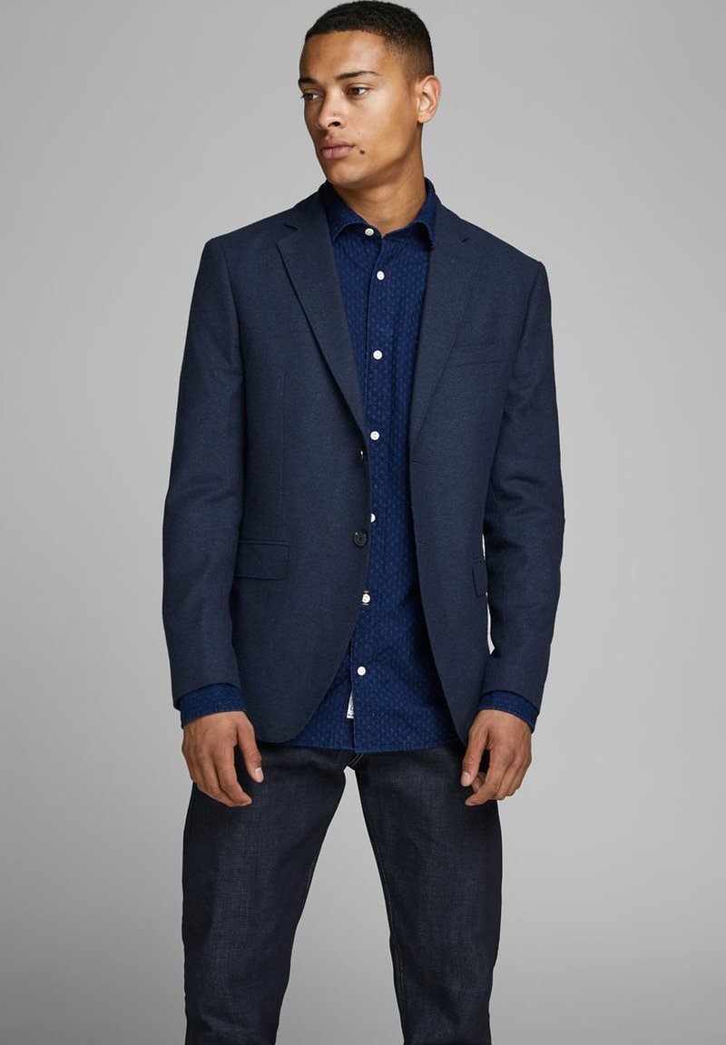 Blazer di lana blu navy con due tasche frontali, indossato sopra una camicia a fantasia blu scuro. Il modello indossa jeans scuri, in piedi contro uno sfondo grigio.
