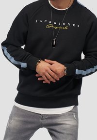 Sudadera de un navy oscuro con acentos en azul claro, que presenta el logo "JACK & JONES Originals" en blanco y amarillo. Tejido texturizado y puños acanalados.