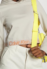 Osoba w beżowej, krótkiej bluzie z kapturem i spodniach z logo Calvin Klein, trzymająca jasnobrązowy pasek torby na ramię w jaskrawym żółtym kolorze.