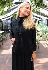 Melna vējjakas veste ar rāvējslēdzēju, ar teksturētu augšdaļu un mīkstu flīsa materiālu, divām priekšējām kabatām, pārlaista pār melnu garām piedurknēm ar spīdumiem.