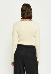 Gerippter, langärmeliger Pullover in Creme mit einem schwarzen gewellten Ausschnitt, kombiniert mit hochtaillierten schwarzen Hosen mit seitlichen Taschen.