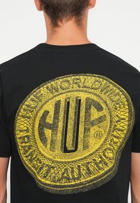 Black cotton t-shirt featuring a large yellow graphic on the back with "HUF" and circular text in a textured design.