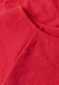 Tissu en coton rouge avec une texture lisse, présentant un col arrondi et un design superposé. Des coutures visibles sont alignées sur les bords.