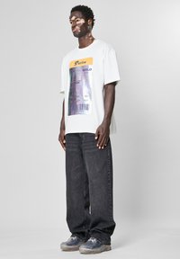 Hvit grafisk t-skjorte med design av flybillett, svarte jeans og gjennomsiktige joggesko. T-skjorten har et holografisk trykk.