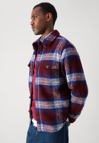 Red and blue plaid flannel shirt with two chest pockets, button closure, and a soft texture, layered over a maroon shirt.