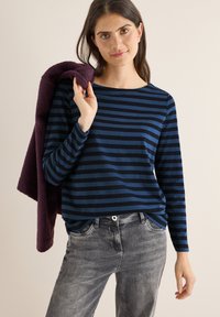 Marineblå og mørkeblå stribet langærmet skjorte med rund halsudskæring, parret med grå denimjeans. Modellen holder en lilla genstand over sin skulder.