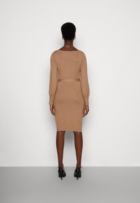 Nainen seisoo valkoista seinää vasten päällään tiukka, polvipituinen beige mekko, jossa on pitkät hihat, sekä mustat korkokengät.