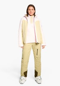 Ski jacket features soft pink and pale yellow panels with a high collar; paired with light yellow pants and white ski boots with minimal detailing.