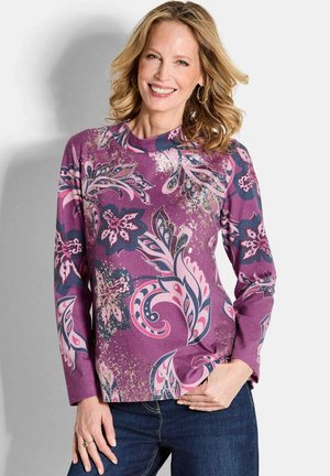 Eine Frau lächelt und trägt ein lila Langarmoberteil mit großen rosa und marineblauen Blumenmustern, blaue Jeans und goldene Creolen.
