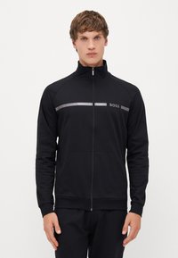 Zwarte zip-up trainingsjacket gemaakt van gladde stof. Heeft een hoge kraag, zijzakken en een zilveren streep met het "BOSS" logo over de borst.