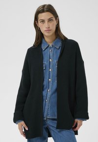 Frau trägt ein blaues Denim-Hemd und Jeans mit einer schwarzen, gerippten Strickjacke, die offen steht, und steht vor einem schlichten hellen Hintergrund.