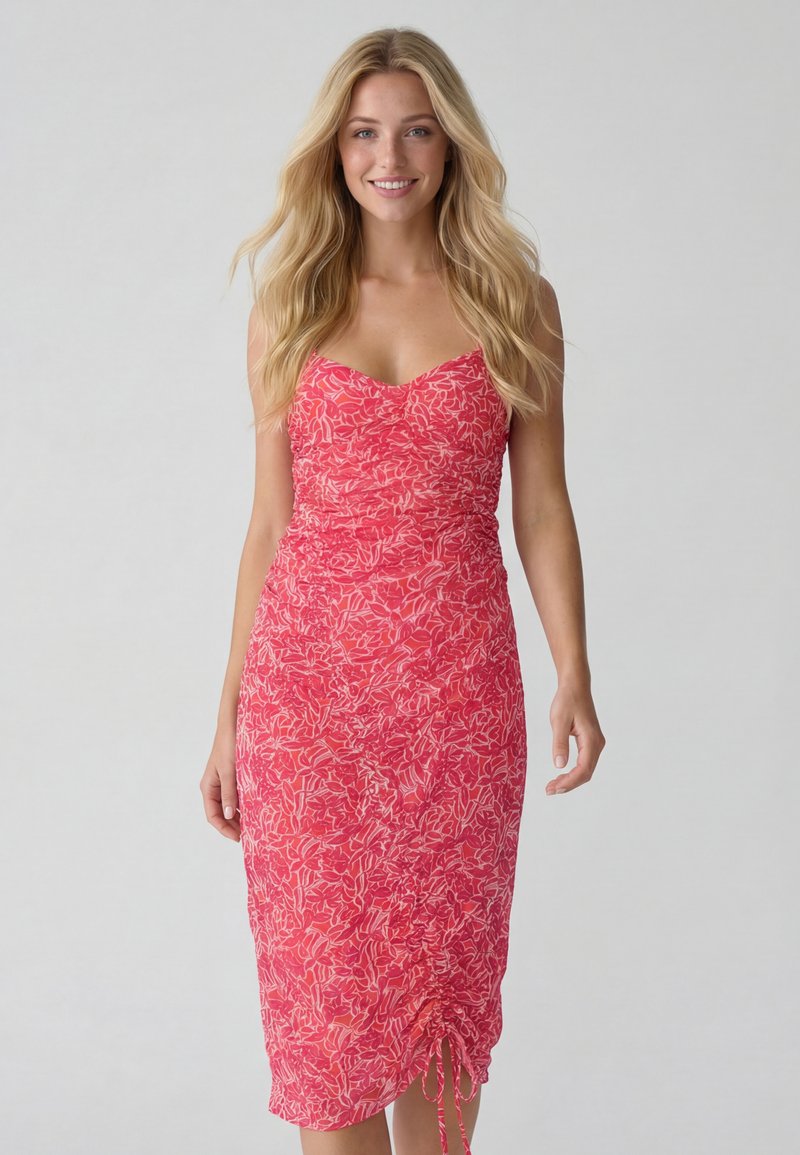 Femme souriante aux longs cheveux blonds portant une robe midi rouge à fleurs cintrée avec un ourlet froncé ajustable à cordon, debout devant un fond uni.
