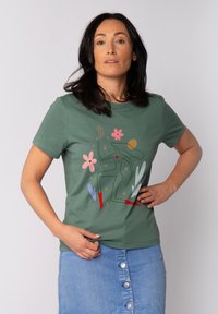 Grünes Baumwoll-T-Shirt mit einem bunten abstrakten Design, das Blumen, eine Figur und Sterne zeigt. Kombiniert mit einem Jeansrock mit Knopfdetails.
