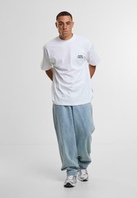 Camiseta blanca de algodón con un pequeño logo negro en el lado izquierdo del pecho, combinada con pantalones de mezclilla azul claro y zapatillas deportivas plateadas.