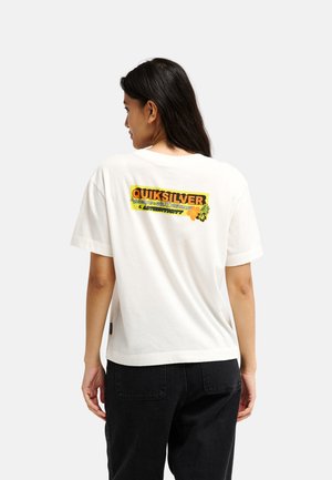 Jeune femme aux longs cheveux foncés portant un t-shirt blanc Quiksilver avec un logo coloré au dos et un pantalon noir, tournée de dos.