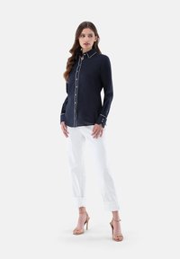 Marineblaues Hemd mit weißen Paspeln, langen Ärmeln und Manschetten. Kombiniert mit weißen hochgekrempelten Hosen und nudefarbenen High Heels.