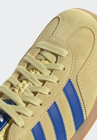 Semišové tenisky v jasně žluté barvě s třemi modrými koženými pruhy, plochými žlutými tkaničkami a gumovou podrážkou. Texturovaný povrch a jemné detaily prošívání.