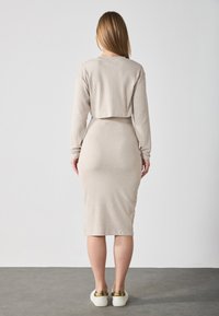 Abito beige a coste con top a maniche lunghe accorciato, silhouette aderente e gonna al ginocchio, abbinato a sneakers bianche con dettagli dorati.