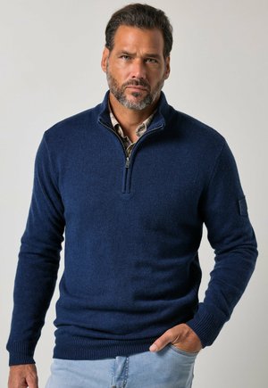 Man met baard die een donkerblauwe sweater met een kwart ritssluiting draagt over een overhemd met kraag, met één hand in de zak van lichtblauwe spijkerbroek, neutrale achtergrond.