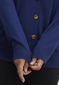 Námorně modrý cardigan se zapínáním na knoflíky, pružnými manžetami a hladkým povrchem, který ukazuje volný střih a jednoduché designové prvky.