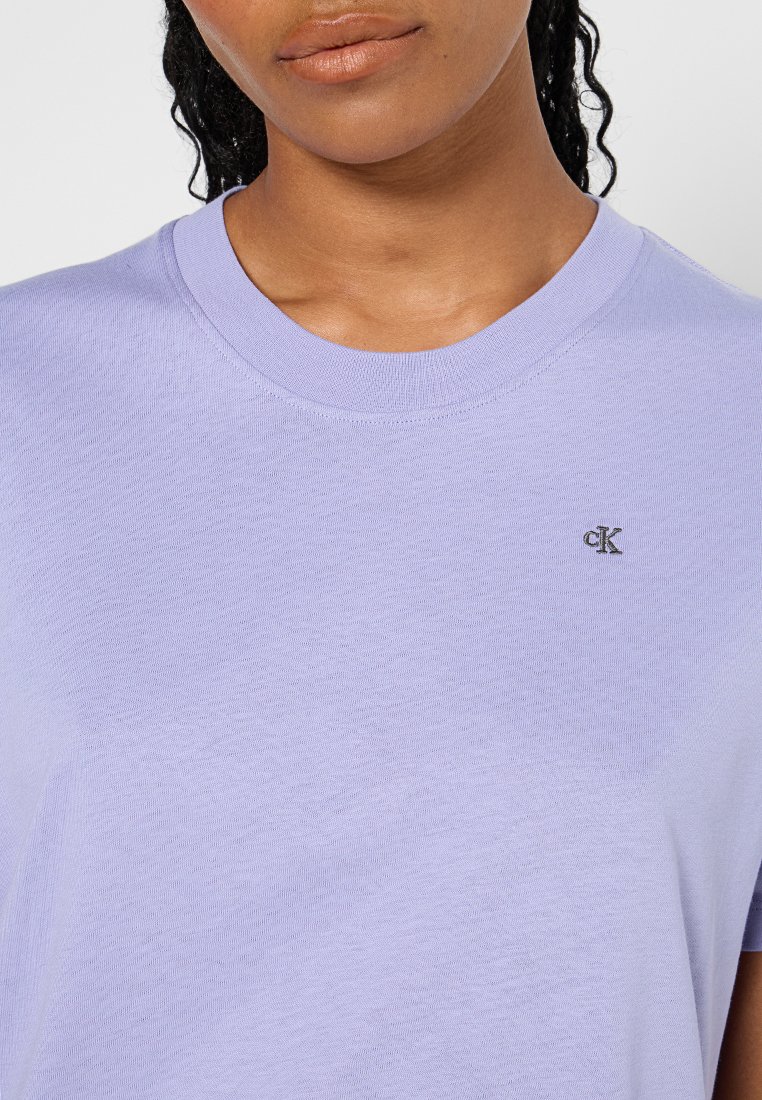 Osoba w jasnofioletowej koszulce z okrągłym dekoltem, na której znajduje się małe, czarne logo "cK" po lewej stronie na klatce piersiowej.