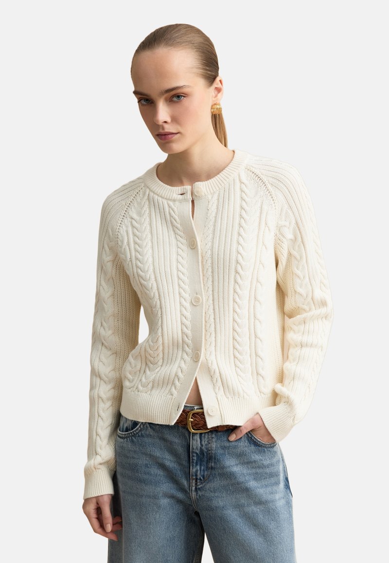 Femme portant un cardigan crème en maille torsadée sur un haut blanc, associé à un jean bleu et une ceinture tressée marron, debout avec une main dans la poche.