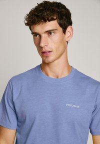 Šviesiai mėlynas medvilninis marškinėliai su trumpomis rankovėmis, su balto siuvinėjimo "PEPE JEANS" logotipu kairėje krūtinės pusėje. Lygus paviršius, apvalus iškirpimas.