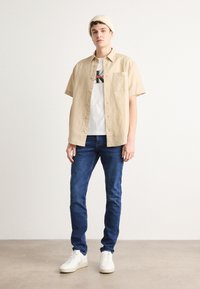 Camicia beige a maniche corte sopra una t-shirt bianca con grafica, abbinata a jeans blu e sneakers bianche, il modello indossa un berretto di cotone beige.