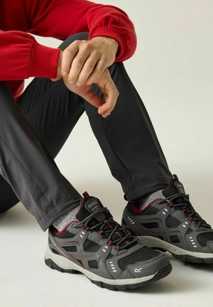 Persoon zittend, draagt zwarte en grijze buitenschoenen, grijze sokken, zwarte broek en een rood shirt met lange mouwen, met handen in elkaar gevouwen over de knie.