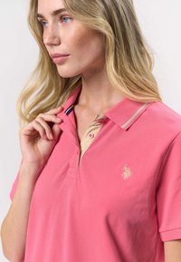 Ružová polokošeľa s béžovým golierom, vybavená dvojtlačidlovým zapínaním, vyšitou značkou a kontrastnými pruhovanými detailmi na golieri.