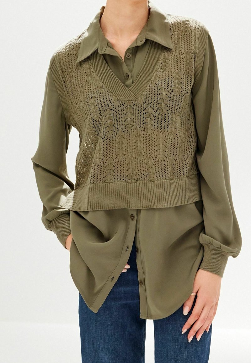 Femme portant une chemise kaki boutonnée avec un gilet tricoté texturé par-dessus et un jean bleu, mains dans les poches et sur le côté.