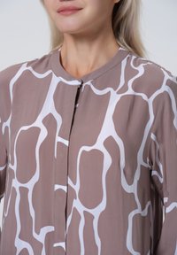 Lange mouwen blouse van een gladde stof, met een taupekleurige basis en een wit abstract patroon, ronde halslijn en sluiting met knopen aan de voorkant.