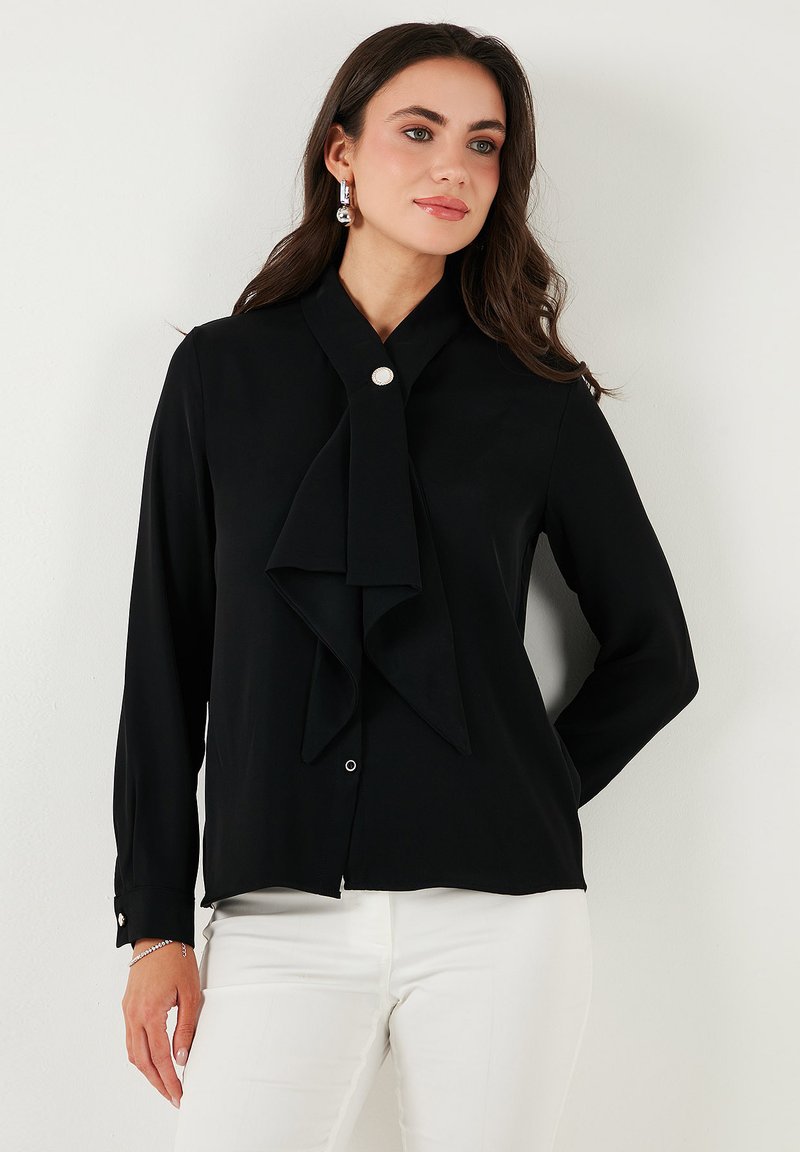 Black long-sleeve blouse with a bow tie detail at the collar, featuring a button closure and white accents. Smooth texture.